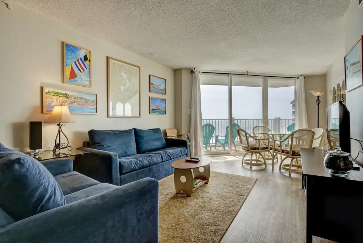 A seamless view of the living and dining areas combined, with blue sofas, a dining table and chairs, large sliding glass doors leading to a balcony, and plenty of natural light streaming in. The room feels open and airy, perfect for socializing and soaking in sunrise views.
