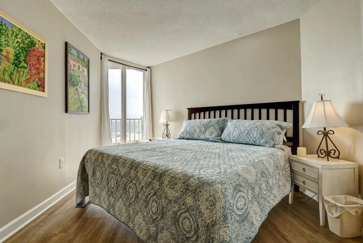 A serene bedroom featuring a bed with a patterned quilt, a nightstand with a lamp, framed artwork on the wall, and large windows letting in natural light. The room feels tranquil and comfortable, perfect for relaxation and rest.