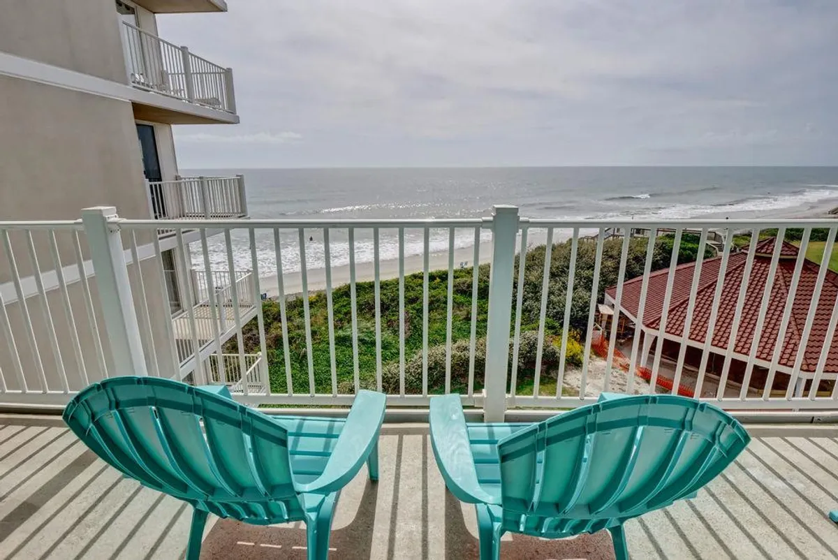 The hero image showcases a stunning, tranquil exterior view from the condo’s balcony. Two inviting turquoise chairs face the endless blue ocean beyond lush green landscaping, with the iconic red-roofed St Regis building nearby. The scene exudes relaxation — perfect for savoring morning coffee, soaking in sunrises, and unwinding with loved ones against a breathtaking coastal backdrop.