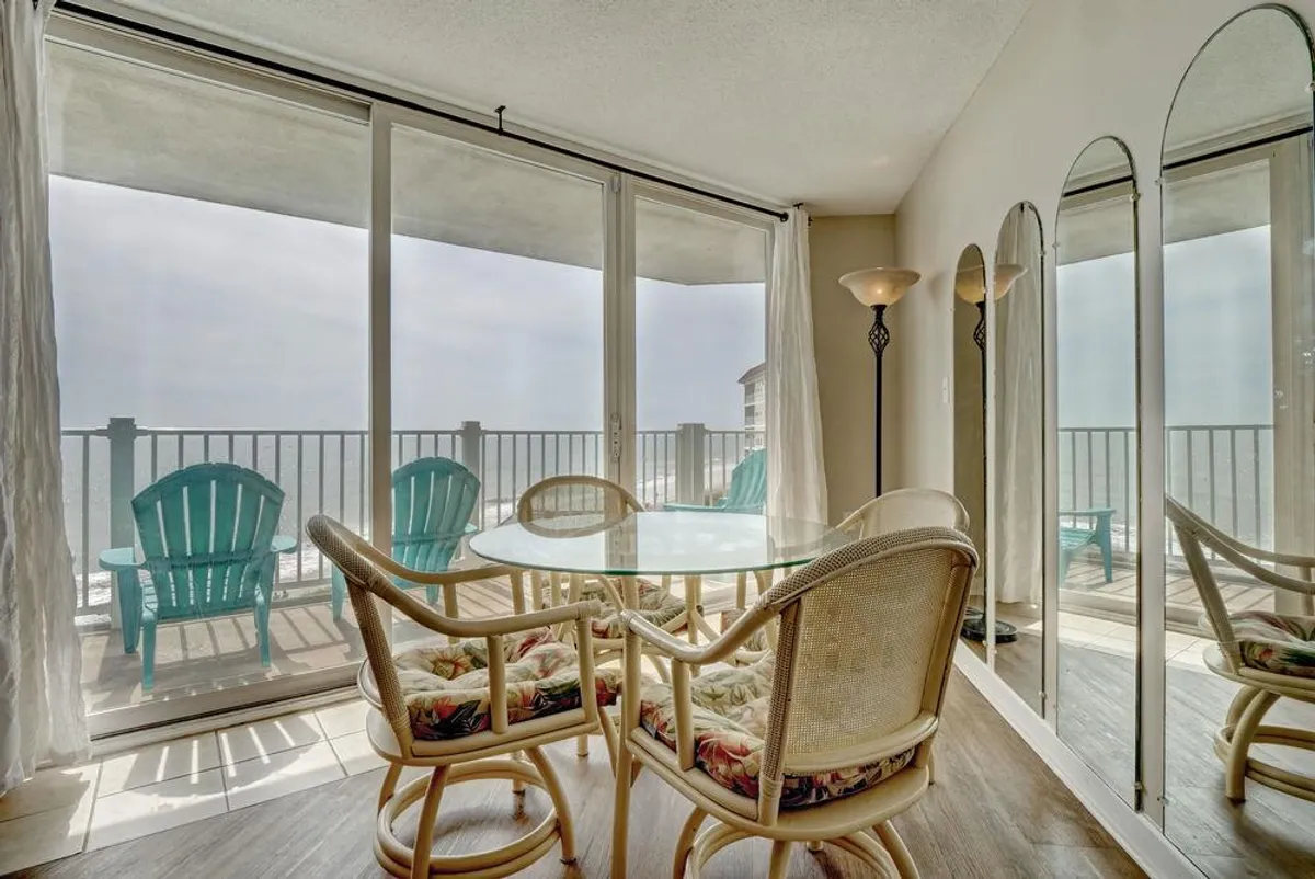 This image showcases a glass dining table surrounded by rattan chairs, positioned near large sliding glass doors that open onto a balcony with sweeping ocean views. The space is flooded with natural light, creating an inviting setting for family meals or morning coffee while enjoying the coastal scenery.