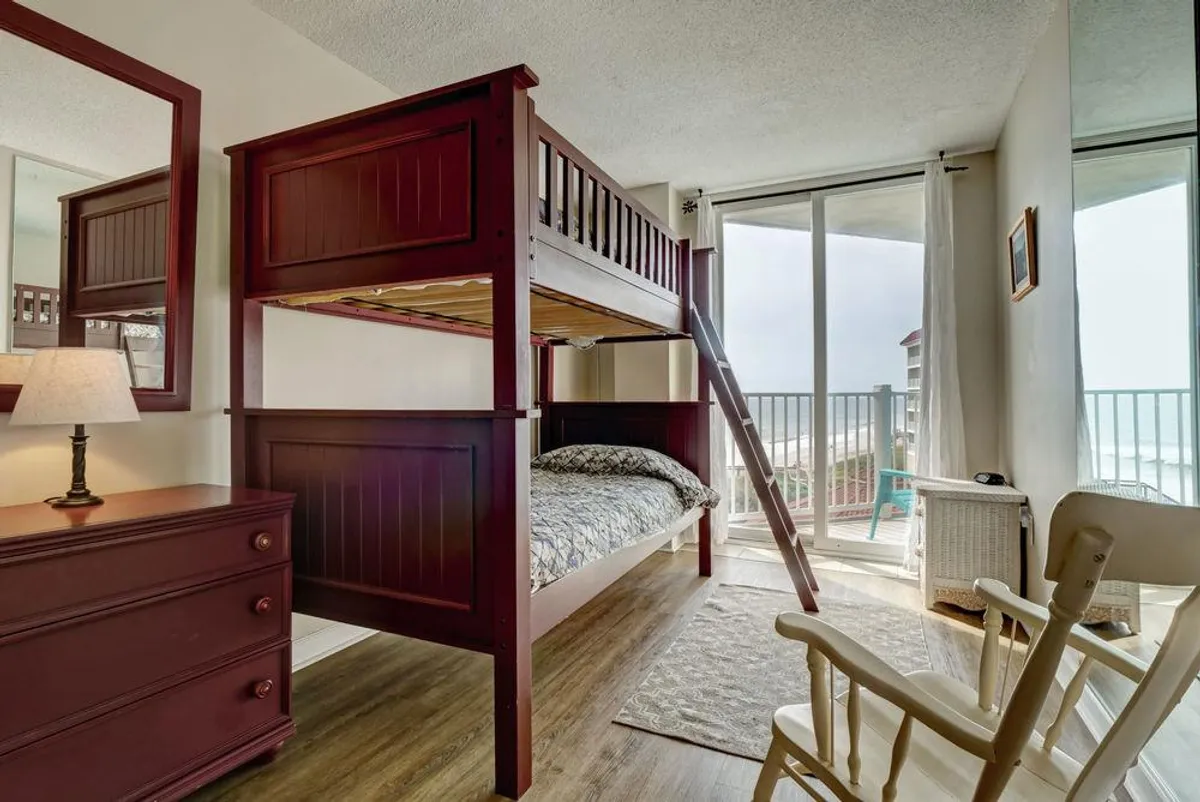 A playful and welcoming bedroom with a twin bunk bed, a rocking chair, a dresser with a lamp, and a large window offering an ocean view. The space feels perfect for children or extra guests, with a relaxed and cheerful mood.