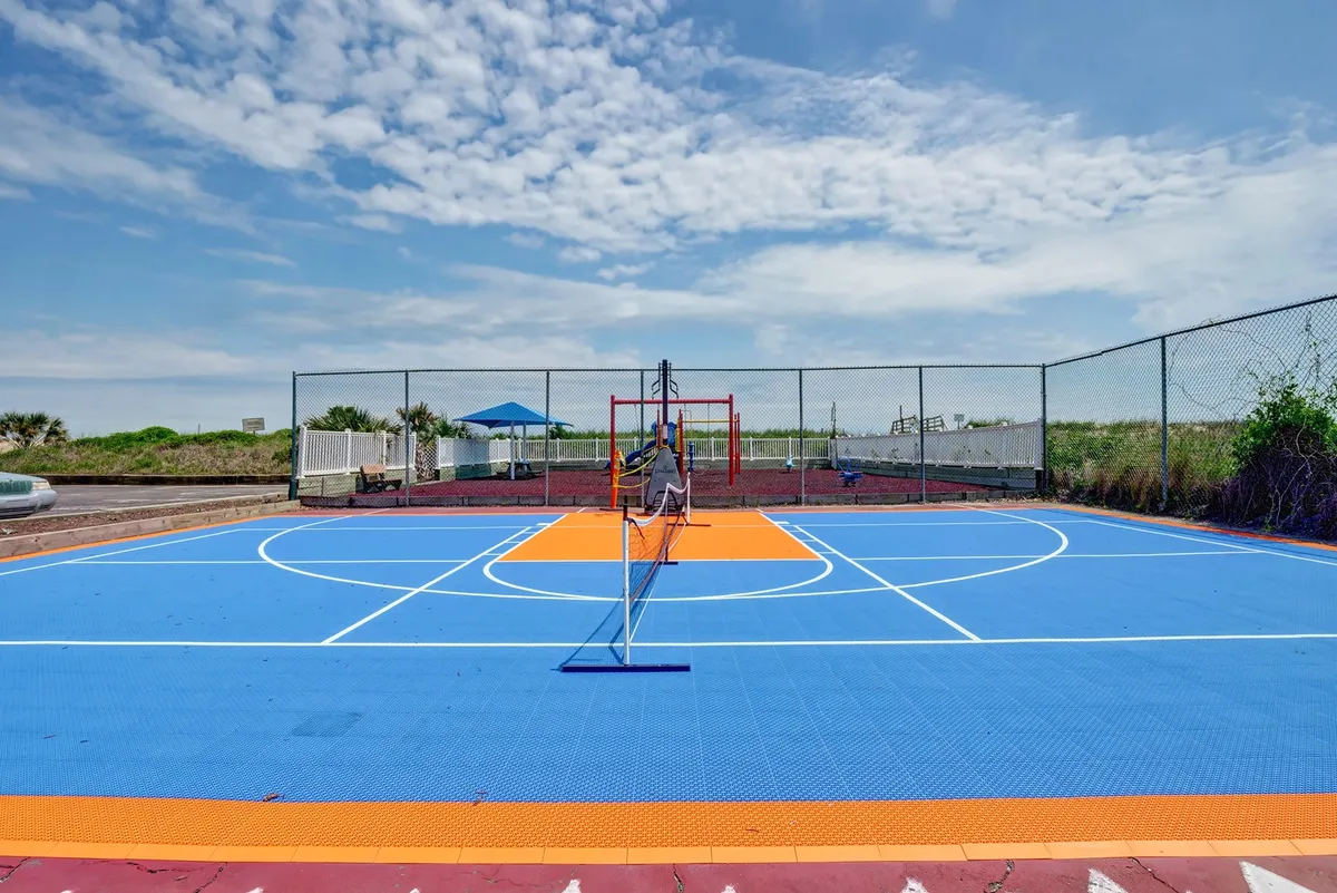 A vibrant exterior view of a colorful basketball court, tennis net, and modern playground equipment beneath a shade structure. The scene is filled with energy, inviting both kids and adults to enjoy sports and play in a safe and cheerful environment.