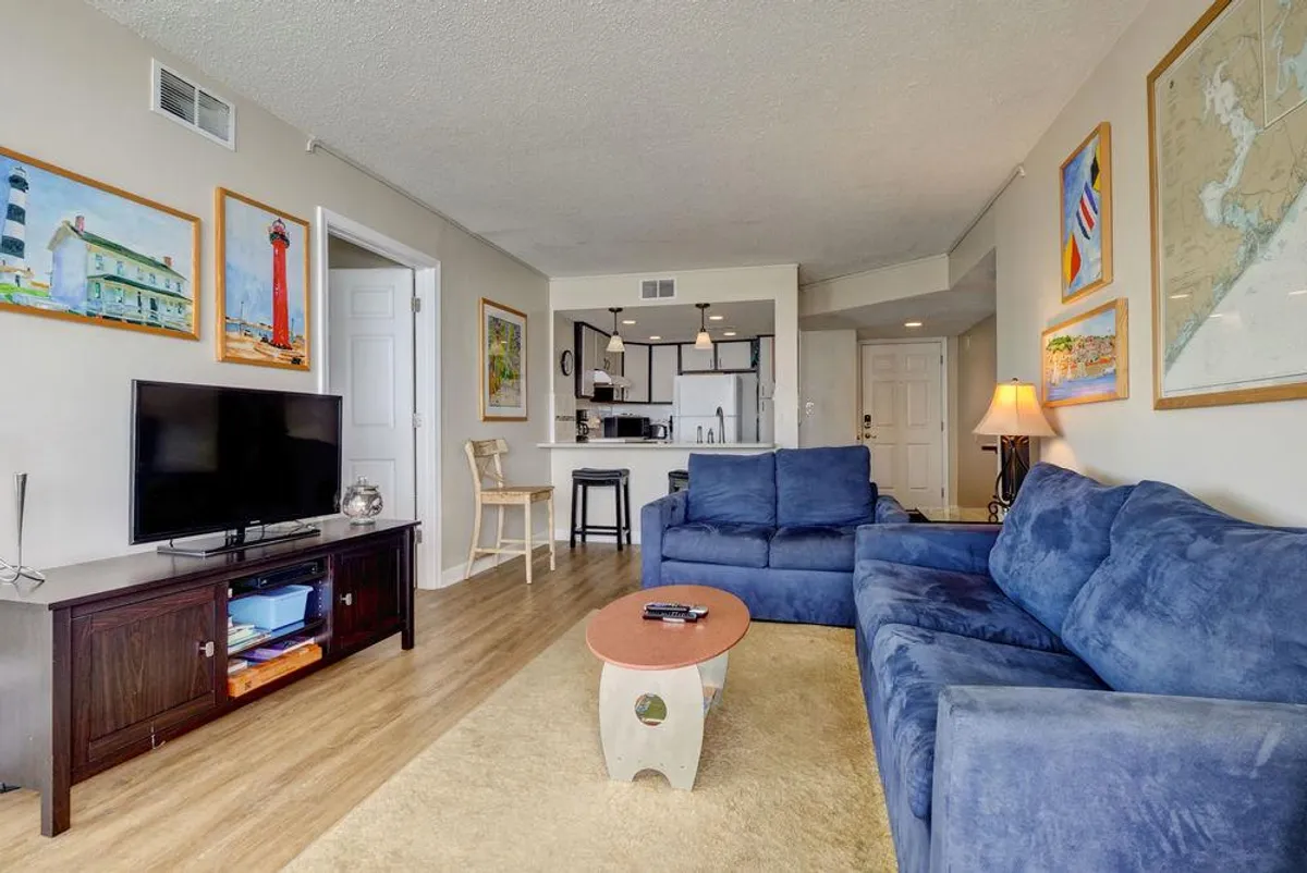 A bright living room featuring a spacious blue sectional sofa, a wooden coffee table set for games or snacks, a television for movie nights, and artwork adorning the walls. This comfortable lounge area is ideal for relaxing with family, watching movies, or enjoying board games.