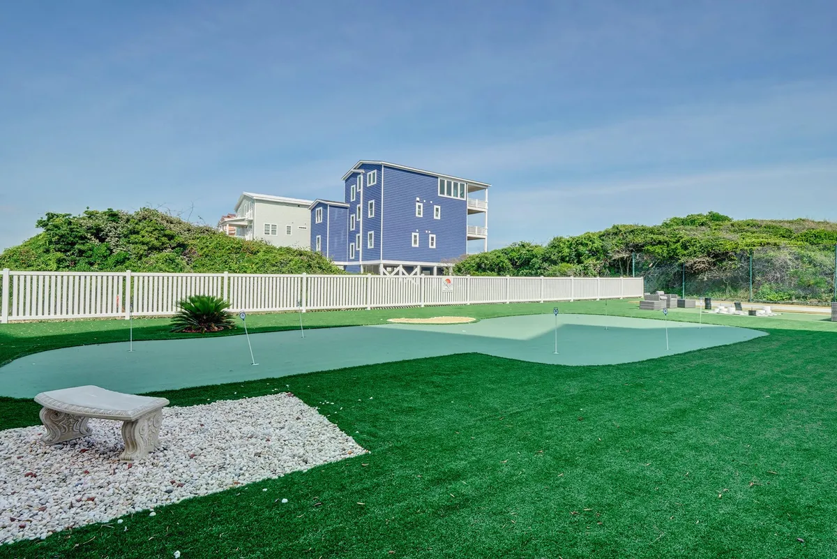 A vibrant outdoor putting green with a decorative stone bench, surrounded by lush greenery and a blue house in the background. The scene is peaceful, with space for both play and quiet reflection, and the bench offers a spot to unwind or chat with friends.